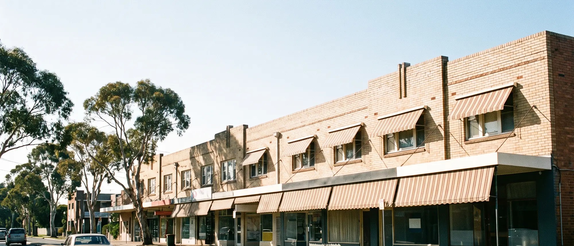 An Australian suburban high street of small shopfronts under awnings in morning sun with gum trees and a wide sky above