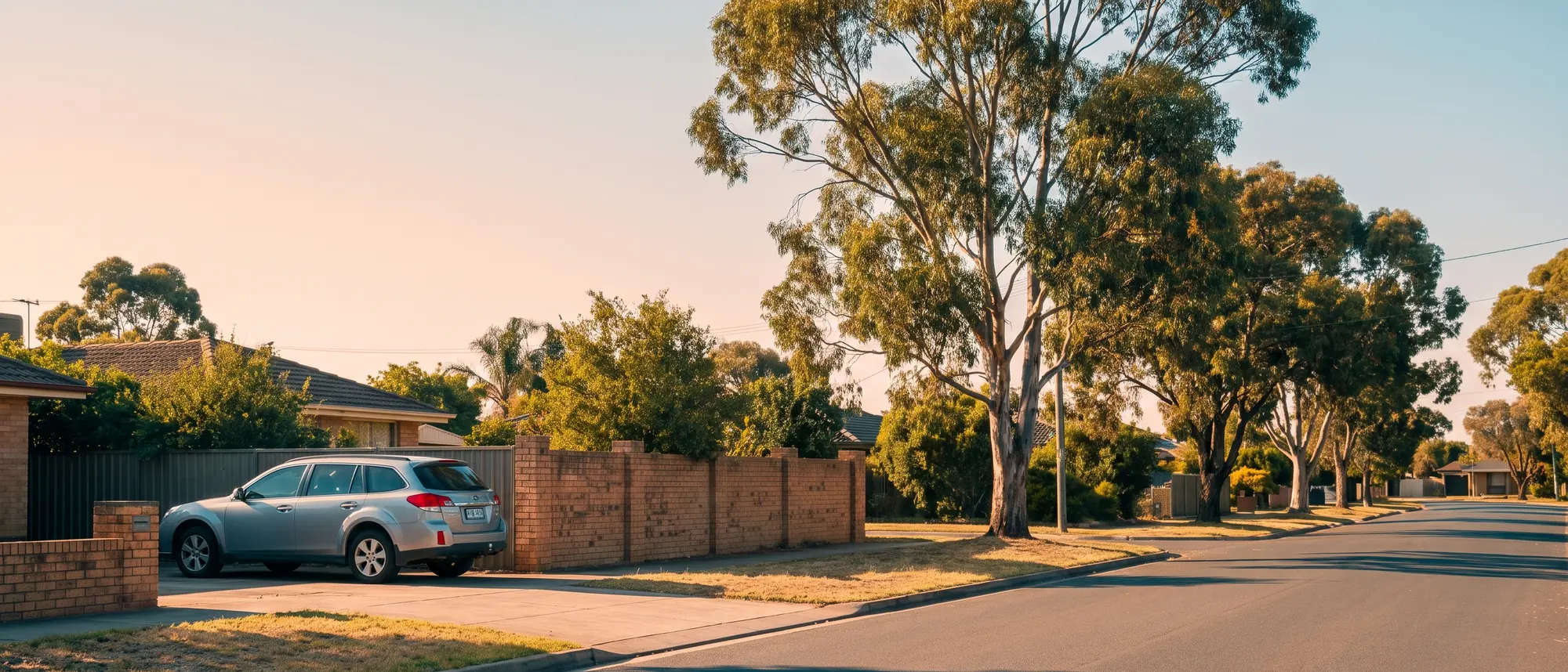 A quiet Australian suburban street in the late afternoon with a car in a concrete driveway and gum trees along the kerb